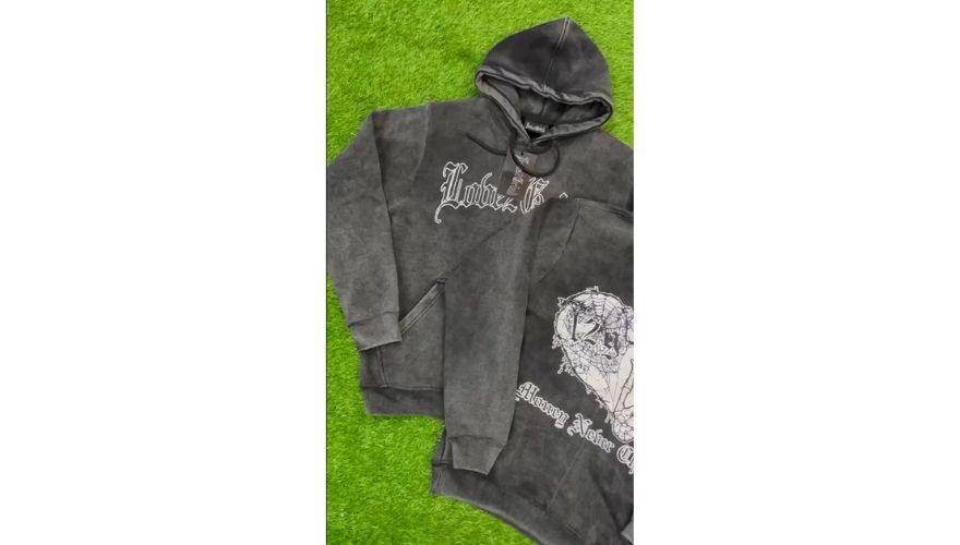 Black acid wash hoodies laid flat on green artificial grass, featuring a faded charcoal finish, gothic style white chest logo on the hoodie, kangaroo pocket, drawstring hood, and matching sweatpants with a large white graphic screen print on the chest, giving a distressed vintage streetwear look.